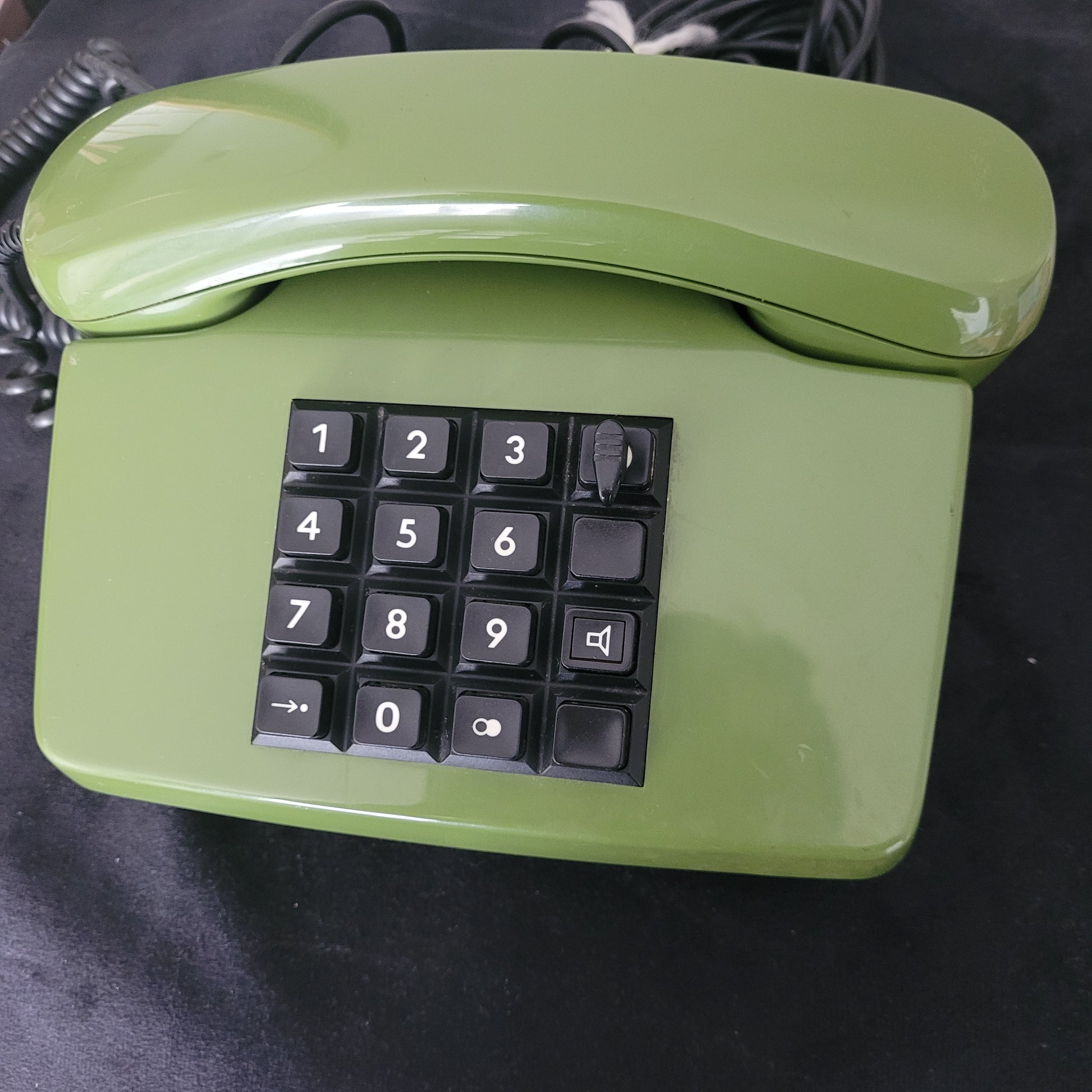 Das Post FetAp 82-2 mit Schlüssel Vintage-Tastentelefon aus den 80er Jahren präsentiert sich in klassischem Grün auf dunklem Hintergrund. Die Vorderseite zeigt die rechteckige Tastatur mit den Zahlen 0 bis 9 sowie zusätzlichen Funktionstasten. Der Hörer liegt links auf der Basisstation und ist mit einem spiralförmigen Kabel verbunden. Ein authentisches Retro-Telefon aus der Telekommunikationsgeschichte – perfekt für Sammler und Liebhaber von Vintage-Technik.