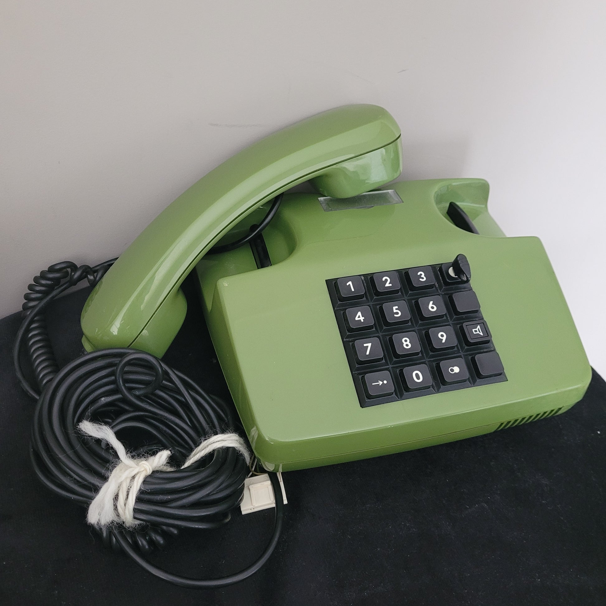 Das Post FetAp 82-2 mit Schlüssel Vintage-Tastentelefon aus den 80er Jahren liegt leicht schräg auf einer schwarzen Oberfläche. Die grüne Farbe und das klassische Design unterstreichen den Retro-Charme. Die rechteckige Form sowie die Tastatur mit den Ziffern 0 bis 9 und zusätzlichen Funktionstasten sind deutlich sichtbar. Das aufgewickelte Kabel ist mit einer weißen Schnur gebunden – ein stilvolles Sammlerstück aus der deutschen Telekommunikationsgeschichte.