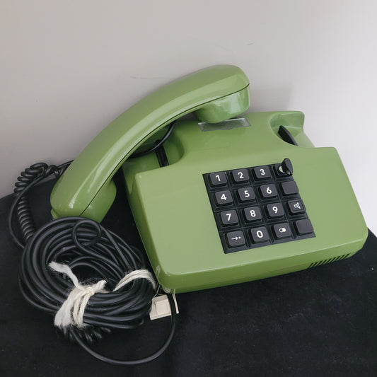 Das Post FetAp 82-2 mit Schlüssel Vintage-Tastentelefon aus den 80er Jahren liegt leicht schräg auf einer schwarzen Oberfläche. Die grüne Farbe und das klassische Design unterstreichen den Retro-Charme. Die rechteckige Form sowie die Tastatur mit den Ziffern 0 bis 9 und zusätzlichen Funktionstasten sind deutlich sichtbar. Das aufgewickelte Kabel ist mit einer weißen Schnur gebunden – ein stilvolles Sammlerstück aus der deutschen Telekommunikationsgeschichte.