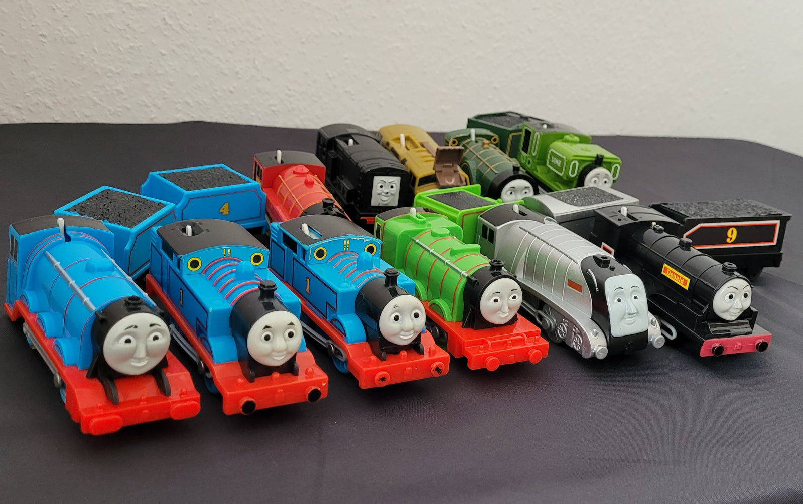 Das Bild zeigt die Vorderseite einer Sammlung von Thomas und seine Freunde-Zügen, die sorgfältig auf einem schwarzen Untergrund arrangiert sind. Im Hintergrund hebt sich ein weißer Hintergrund ab, wodurch die verschiedenen Lokomotiven besonders gut zur Geltung kommen.