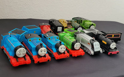 Das Bild zeigt die Vorderseite einer Sammlung von Thomas und seine Freunde-Zügen, die sorgfältig auf einem schwarzen Untergrund arrangiert sind. Im Hintergrund hebt sich ein weißer Hintergrund ab, wodurch die verschiedenen Lokomotiven besonders gut zur Geltung kommen.