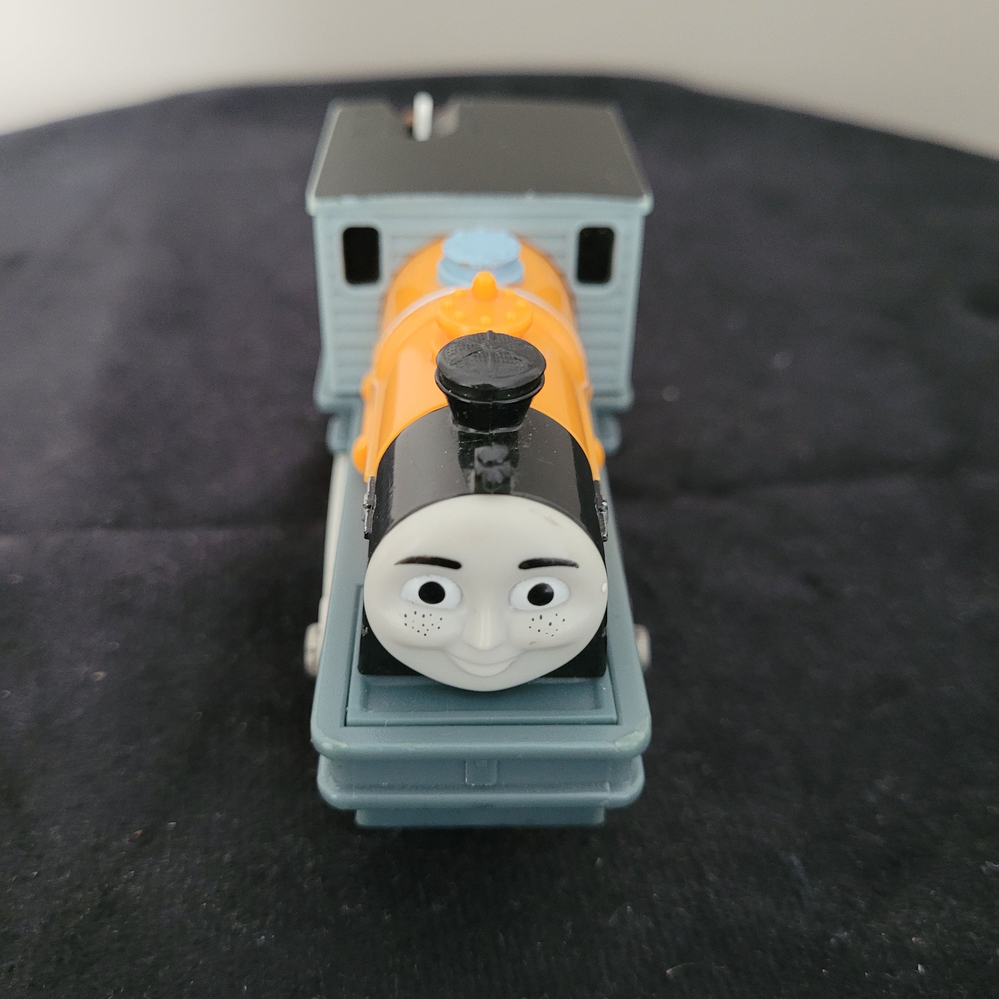 Das Bild zeigt die Lokomotive Dash, Modell 1572WC, auf einem schwarzen Untergrund. Der Zug ist in leuchtendem Gelb und Blau gehalten, mit detaillierten Rädern und einem charakteristischen Gesicht, das an die Figur aus ‚Thomas und seine Freunde‘ erinnert.