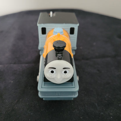 Das Bild zeigt die Lokomotive Dash, Modell 1572WC, auf einem schwarzen Untergrund. Der Zug ist in leuchtendem Gelb und Blau gehalten, mit detaillierten Rädern und einem charakteristischen Gesicht, das an die Figur aus ‚Thomas und seine Freunde‘ erinnert.