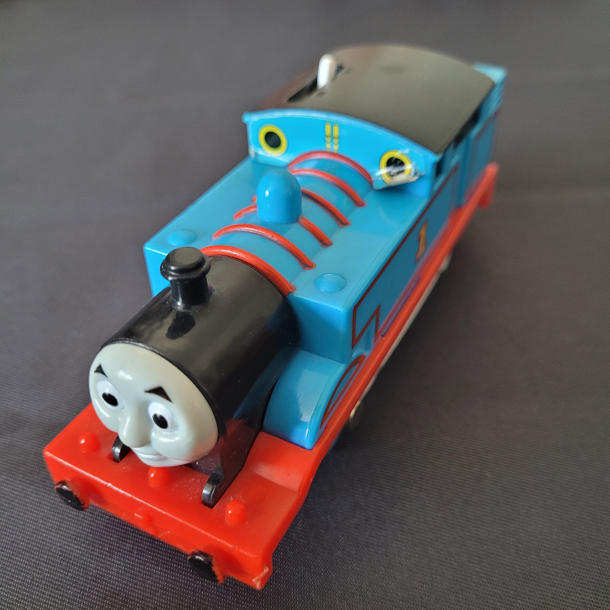 Das Bild zeigt die Lokomotive Thomas, Modell 1840D1, auf einem schwarzen Untergrund. Die blaue Lok mit rotem Fahrgestell und grauen Rädern ist detailliert gestaltet und zeigt die bekannten Gesichtszüge von Thomas aus der Serie ‚Thomas und seine Freunde‘