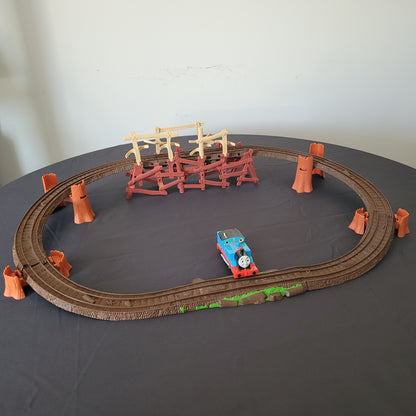 Das vollständige Thomas und seine Freunde Shake, Shake Bridge Playset, bestehend aus der Brücke, den dazugehörigen Schienen und der Lokomotive Thomas. Das Set ist auf einem schwarzen Untergrund arrangiert, während der Hintergrund weiß ist, wodurch die einzelnen Elemente besonders gut zur Geltung kommen.