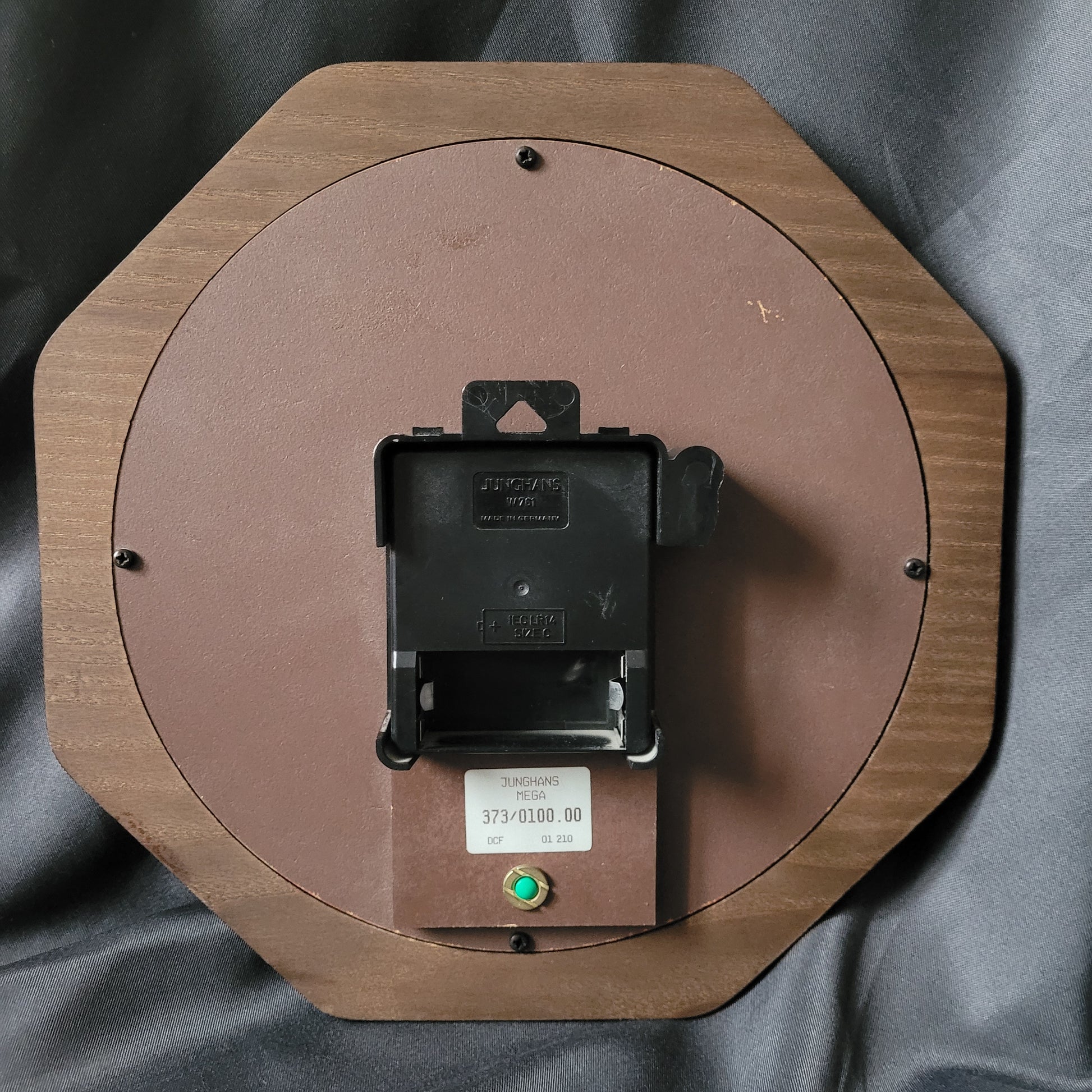 Rückseite der Wanduhr Junghans Mega Radio Controlled Vintage mit achteckigem Holzrahmen, rundem Rückwandpanel und Quarz-Uhrwerk, sichtbarem Batteriefach für eine AA-Batterie, Aufkleber mit Herstellerangaben sowie grünem Bedienelement, klassische Funkuhr im Vintage-Design.
