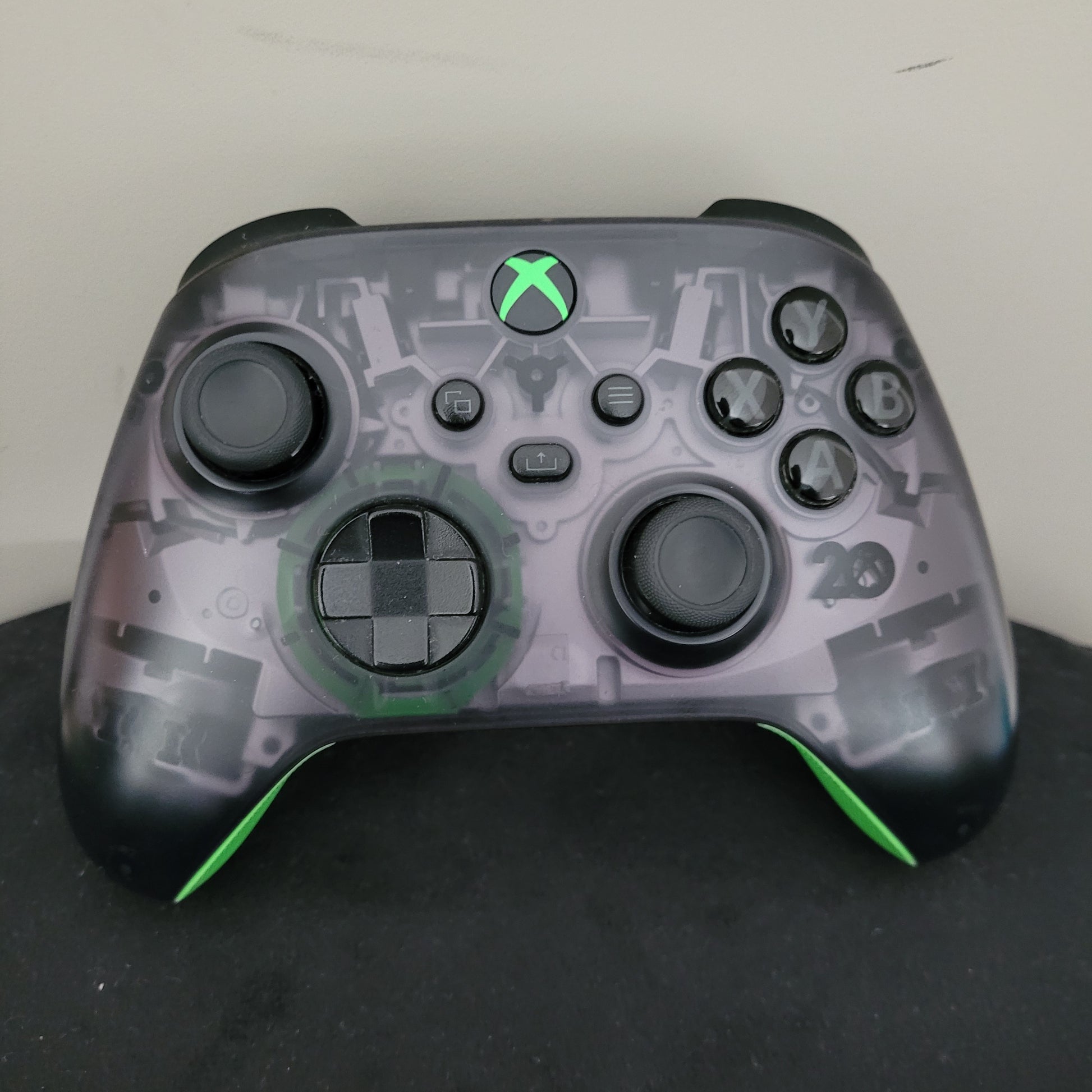 Das Bild zeigt die Vorderansicht des Xbox Wireless Controller – 20th Anniversary Special Edition. Erkennbar sind das transparente, rauchgraue Gehäuse mit sichtbaren Innenkomponenten, schwarze Bedienelemente wie Joysticks, D-Pad und Tasten (A, B, X, Y), sowie die grün leuchtende Xbox-Taste mittig. Die grünen Akzente an den Griffen und die klassische Formgebung unterstreichen den Gaming-Charakter und den Jubiläumsstil des Controllers. Der Zustand wirkt gepflegt.