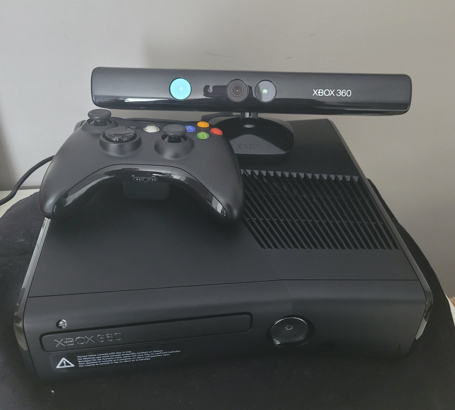 Xbox 360 S Modell 1439 – Konsole mit Zubehör auf schwarzem Untergrund und weißem Hintergrund Das Bild zeigt die Vorderseite einer Xbox 360 S Modell 1439, zusammen mit dem zugehörigen Netzteil, einem Controller und einer Sensorleiste. Die Konsole hat ein schlankes, schwarzes Design und ist auf einem schwarzen Untergrund platziert, während der Hintergrund weiß ist. Die Zubehörteile sind neben der Konsole angeordnet, um einen vollständigen Überblick über das Gaming-Set zu geben.