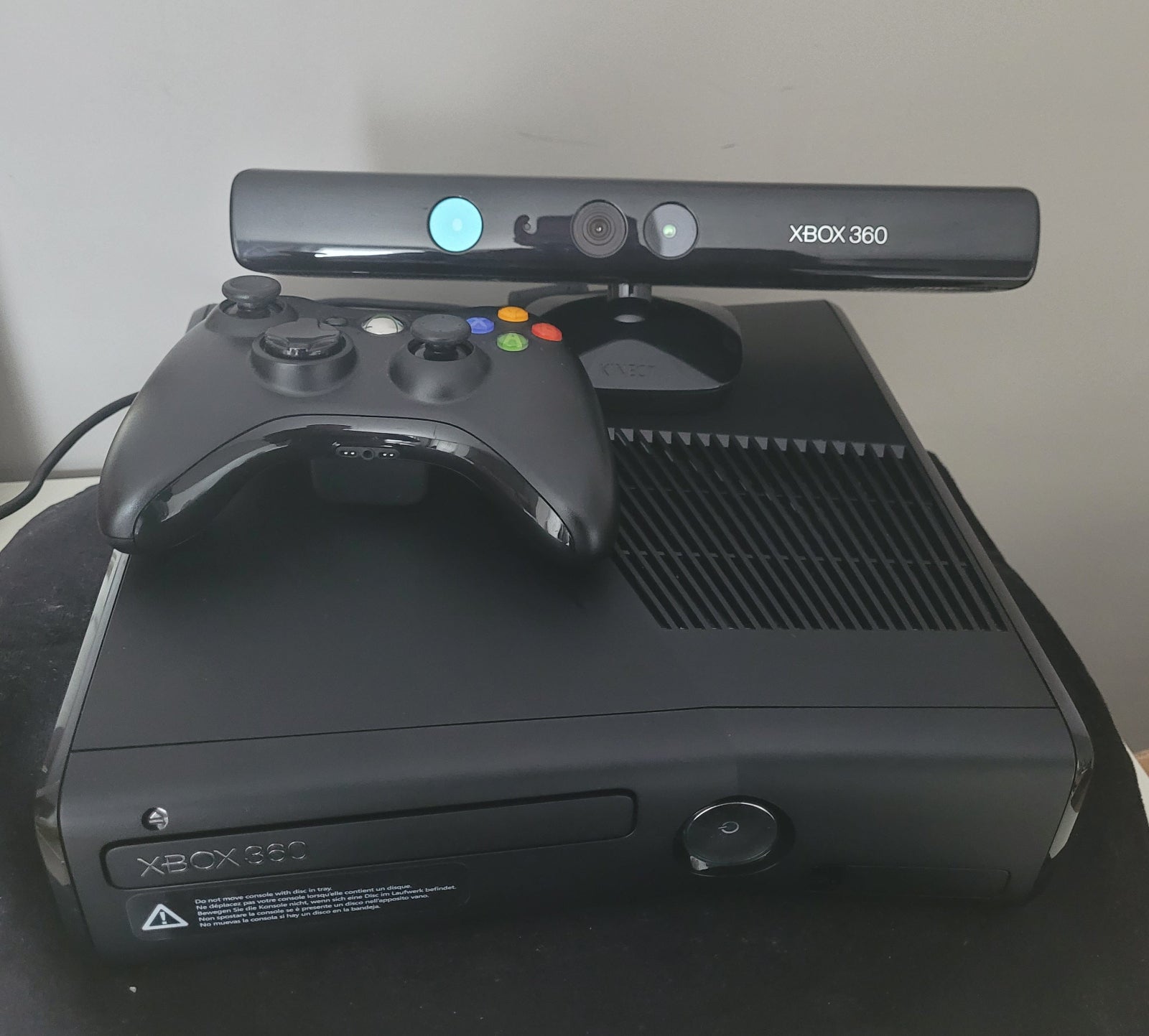 Xbox 360 S Modell 1439 – Konsole mit Zubehör auf schwarzem Untergrund und weißem Hintergrund Das Bild zeigt die Vorderseite einer Xbox 360 S Modell 1439, zusammen mit dem zugehörigen Netzteil, einem Controller und einer Sensorleiste. Die Konsole hat ein schlankes, schwarzes Design und ist auf einem schwarzen Untergrund platziert, während der Hintergrund weiß ist. Die Zubehörteile sind neben der Konsole angeordnet, um einen vollständigen Überblick über das Gaming-Set zu geben.