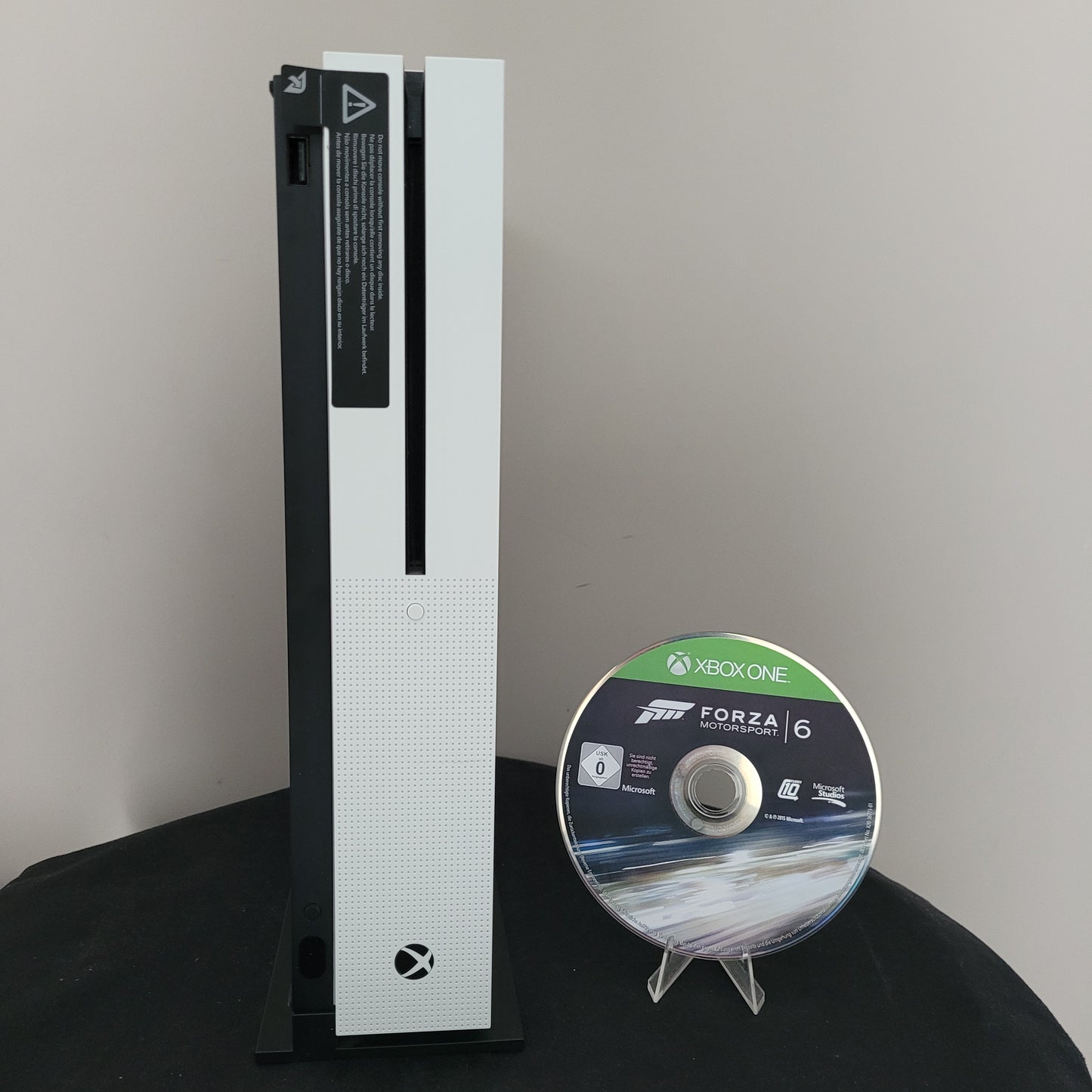 Die Vorderseite einer weißen Xbox One S Konsole ist deutlich zu erkennen. Sie zeigt das eingelassene Xbox-Logo, einen USB-Port und den Disc-Schlitz mit schwarzem Akzent. Daneben steht die Forza Motorsport 6 Spiel-CD, aufrecht präsentiert, mit sichtbarem Logo und ohne Originalverpackung. Das Arrangement vermittelt einen gepflegten, vintage-inspirierten Zustand des angebotenen Gaming-Bundles.