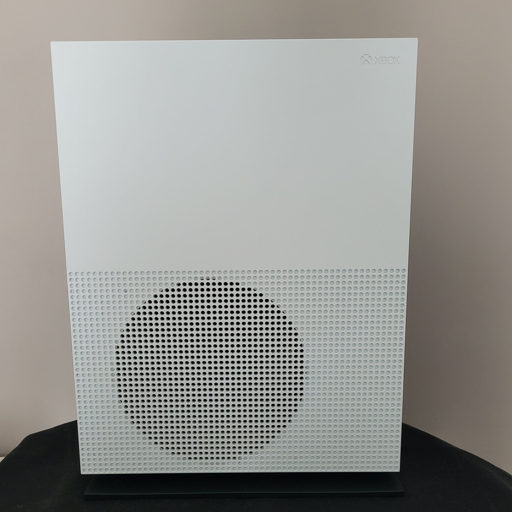 Das Foto zeigt die Seitenansicht einer weißen Xbox One S Konsole in vertikaler Position. Erkennbar ist das charakteristische große, runde Lüftungsgitter mit Wabenstruktur auf der rechten Geräteseite, während das dezente Xbox-Logo links oben eingelassen ist.