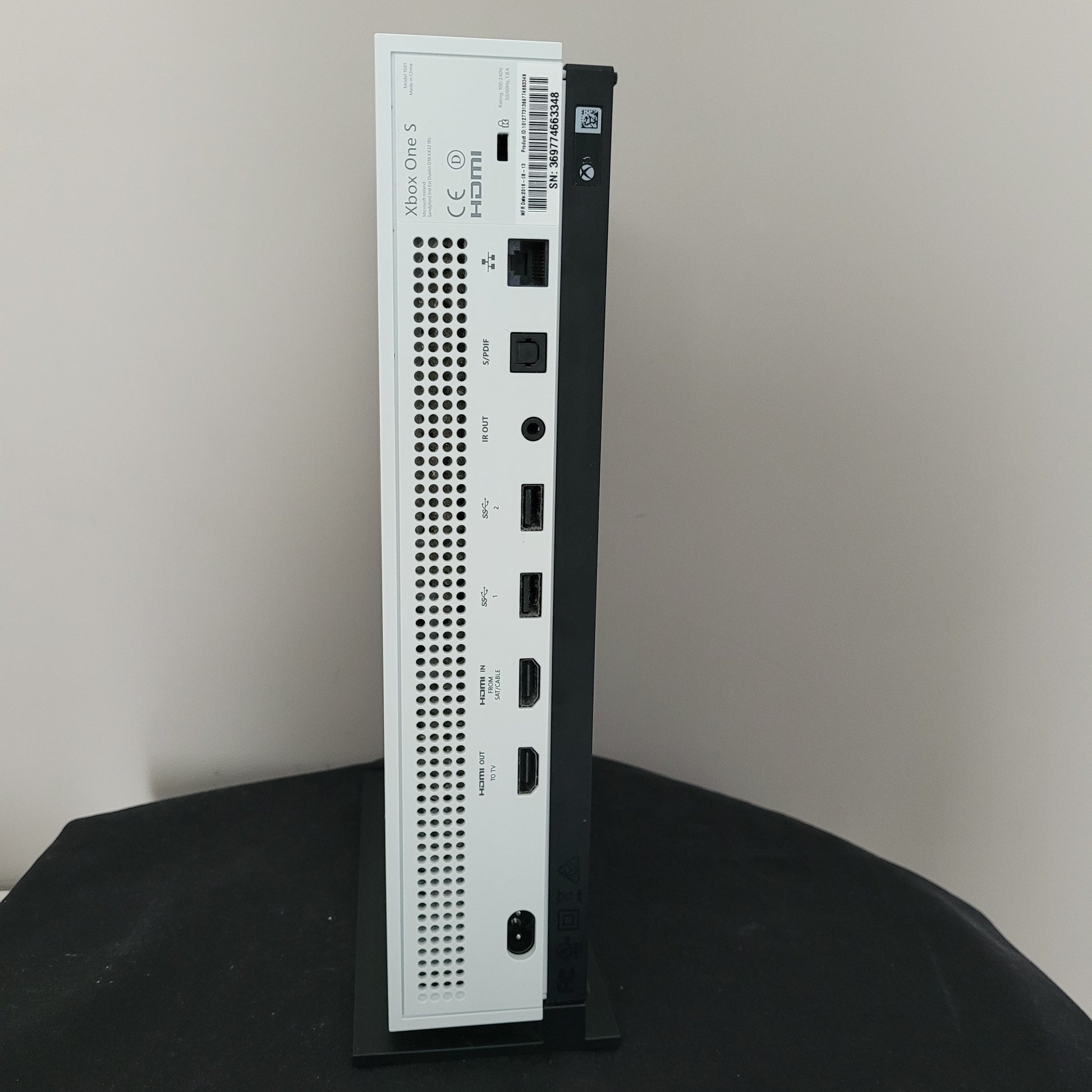 Das Foto zeigt die Rückseite einer weißen Xbox One S Konsole. Sichtbar sind mehrere Anschlüsse in einer horizontalen Reihe: darunter Ethernet, S/PDIF, IR-Out, zwei USB-Ports, HDMI-In, HDMI-Out sowie der Stromanschluss. Über den Ports befindet sich ein Seriennummer-Aufkleber („SN: 369717665346“), darunter ist ein längliches Lüftungsgitter integriert. Links unten sind das Xbox One S Logo, das HDMI-Emblem und diverse Zertifizierungszeichen zu erkennen. 