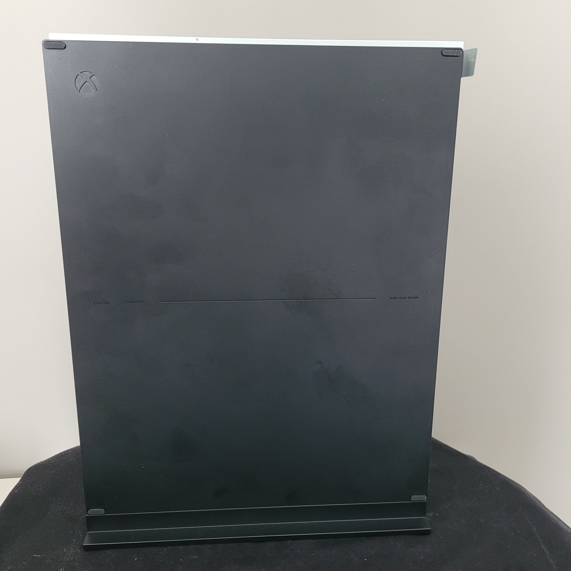 Das Foto zeigt die Seitenansicht einer weißen Xbox One S Konsole in vertikaler Position. Erkennbar ist das charakteristische große, schwarze Oberfläche während das dezente Xbox-Logo links oben eingelassen ist.