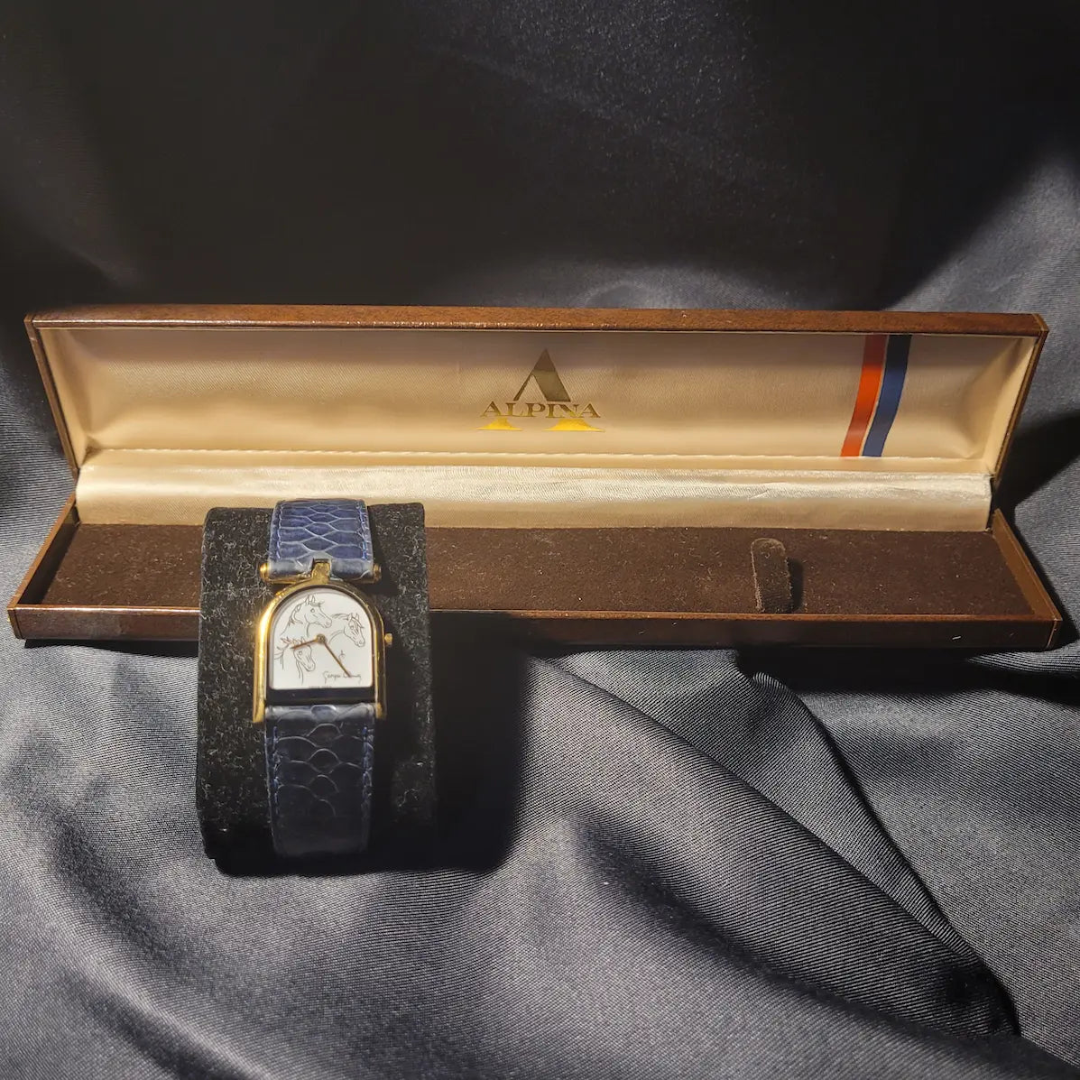 Elegante Alpina Georges Claude Armbanduhr mit drei Pferdeköpfen auf dem weißen Zifferblatt, goldenem Gehäuse und blauem Lederarmband. 