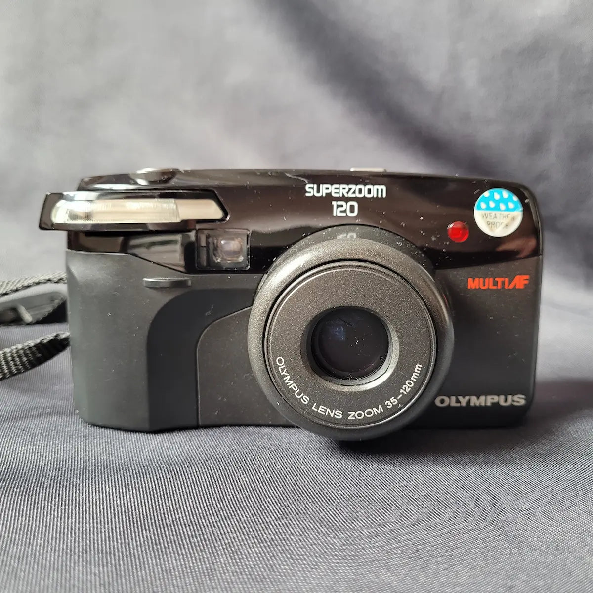 Schwarze Olympus Superzoom 120 Multi AF Analogkamera mit ausgefahrenem Zoom-Objektiv. Frontseite.